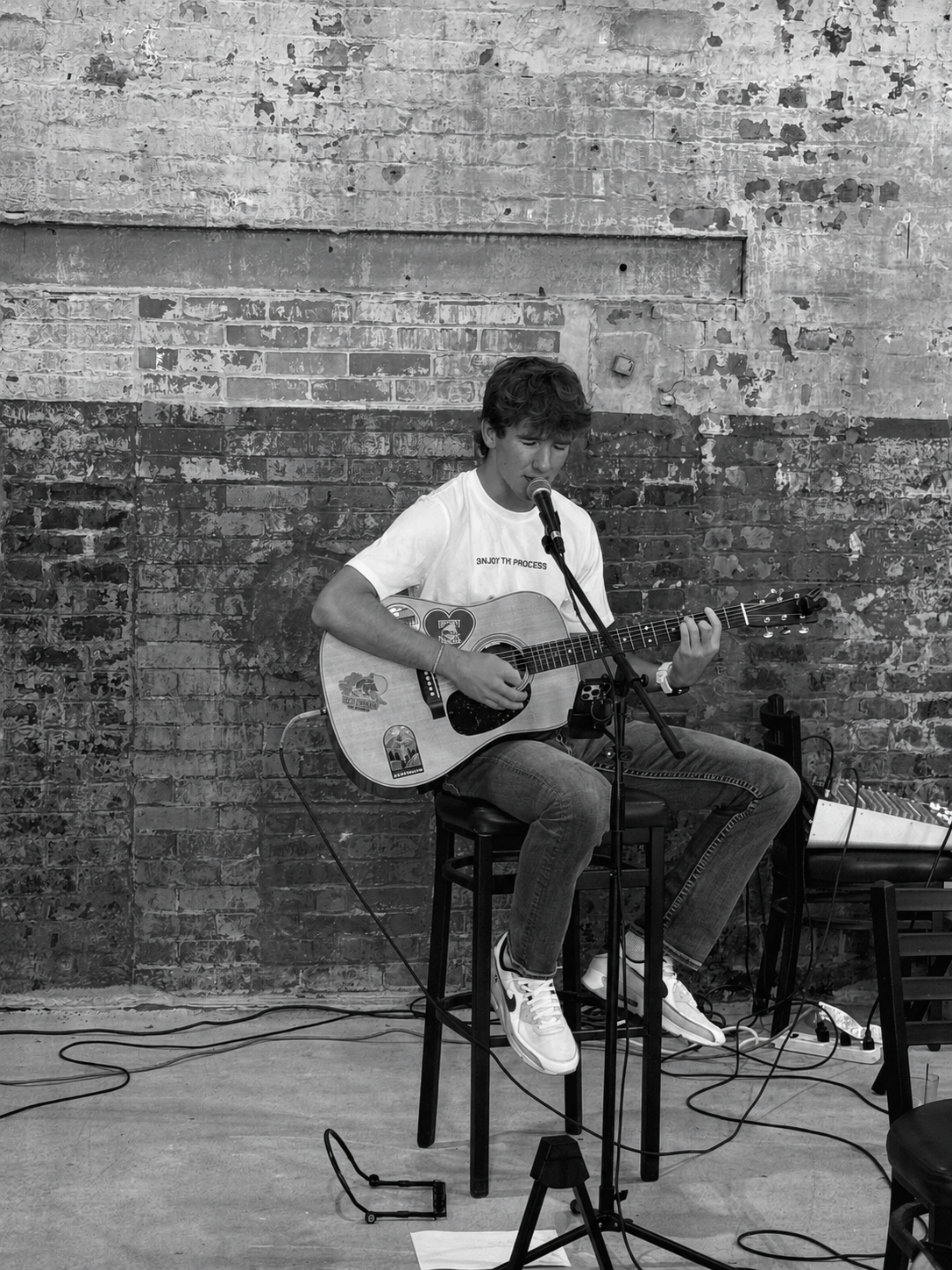 Ryan North seated on a stool playing acoustic guitar against a weathered brick wall